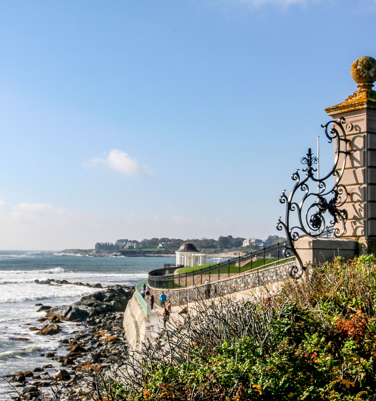 A coastal promenade with ornate stone pillars and wrought-iron gates overlooking the rocky shoreline and calm sea.