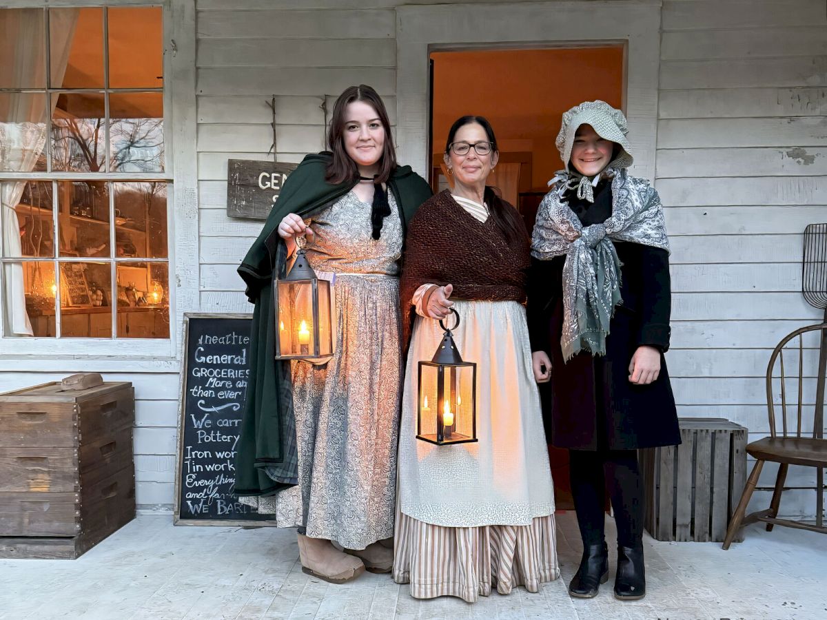 Three people in period clothing holding lanterns stand outside a building, with a chalkboard sign to the side.