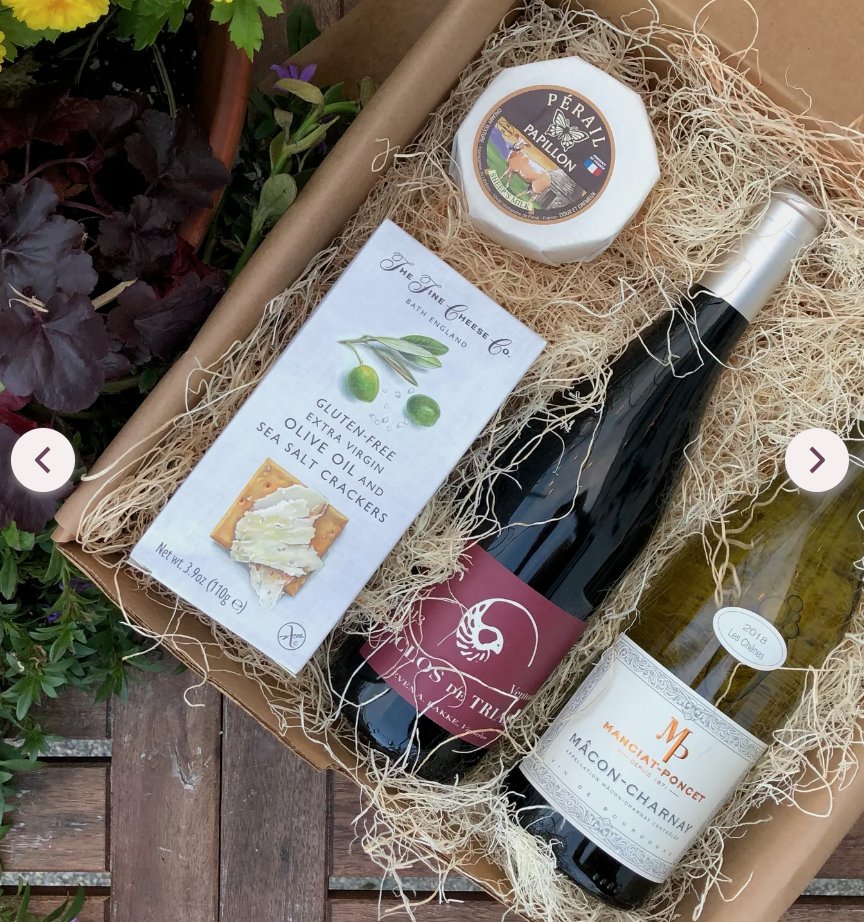 A gift box with wine bottles, a soap bar, a small candle, and a plant-flower backdrop, arranged on shredded filler in a cardboard tray.