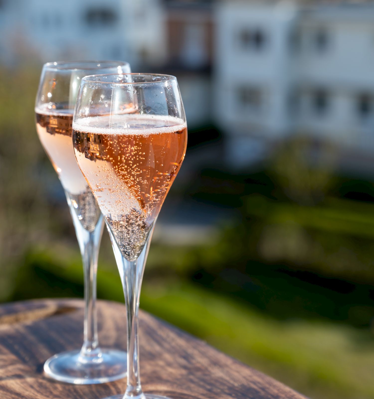 Three champagne flutes filled with rose wine sit on a wooden railing, blurred rooftops in the background.