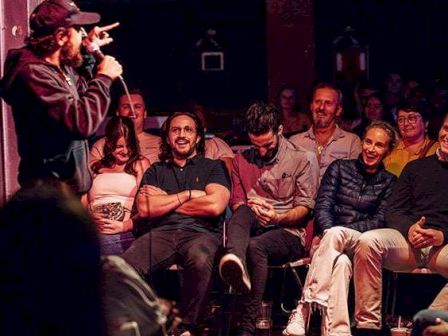 A comedian performs on stage to a seated audience in a small venue, with several people smiling and watching intently.