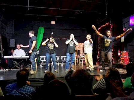 A group of seven performers on a stage with a green curtain, microphones, and an audience watching; one sits at a keyboard left, others sing or pose, colorful lighting in the background.