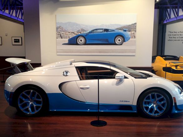 A white and blue Bugatti Veyron on display in a museum with other sports cars and a framed blue car photo in the background.