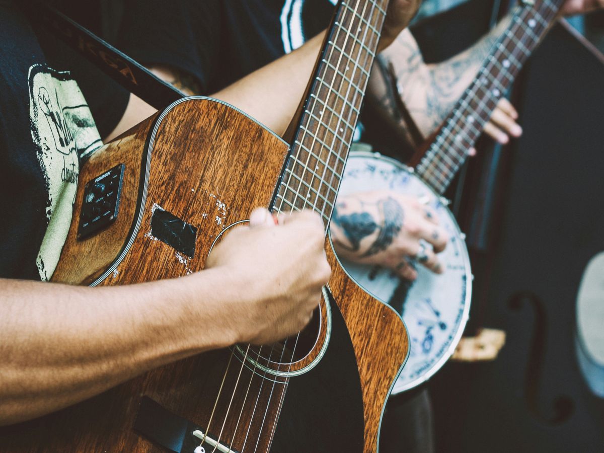 Two people playing acoustic guitars closely, strings and wooden bodies in focus, pick and tattooed arms visible, music in action.