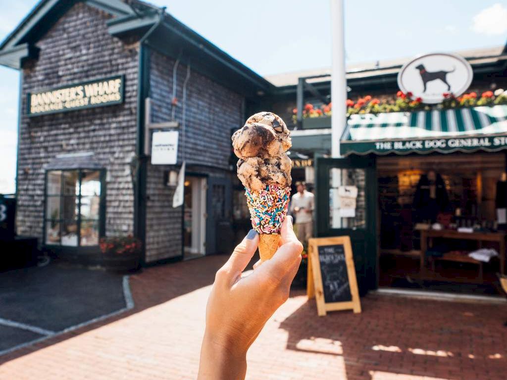 A hand holds a waffle ice-cream cone with chocolate and sprinkles outside a street cafe with signs and flowers in the background.