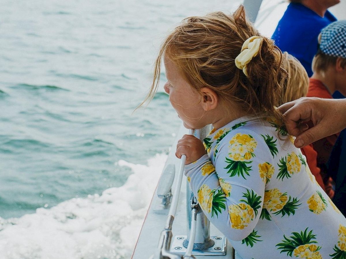 A little girl in a yellow-floral dress on a boat, as an adult helps her while waves roll by.