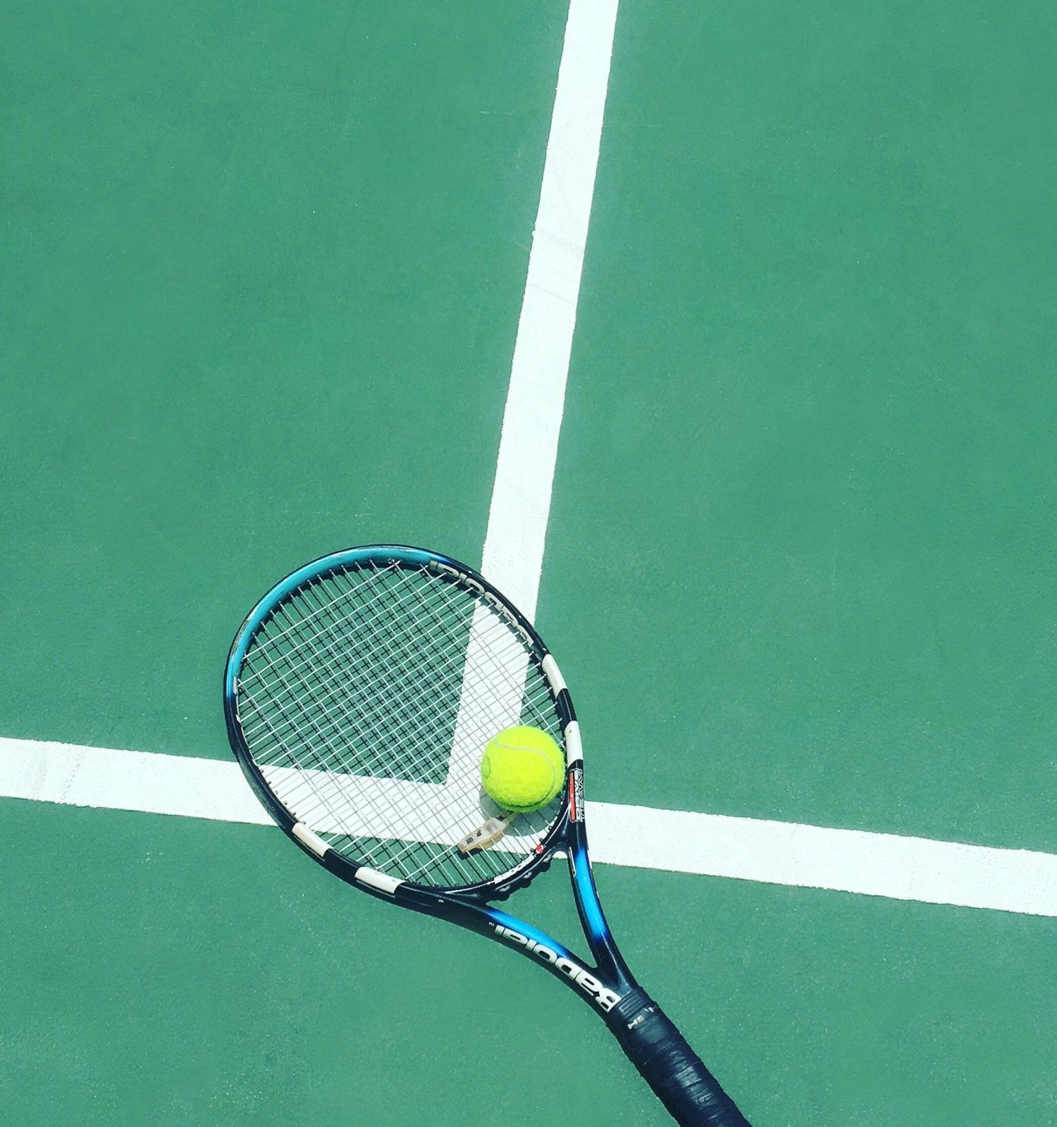 A tennis racket and yellow ball rest on a green court with white lines, ready for the next shot.