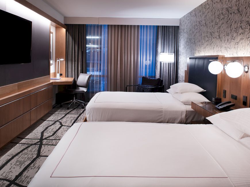 The image shows a modern hotel room with two beds, a TV, desk, and patterned carpet, creating a cozy and stylish atmosphere.
