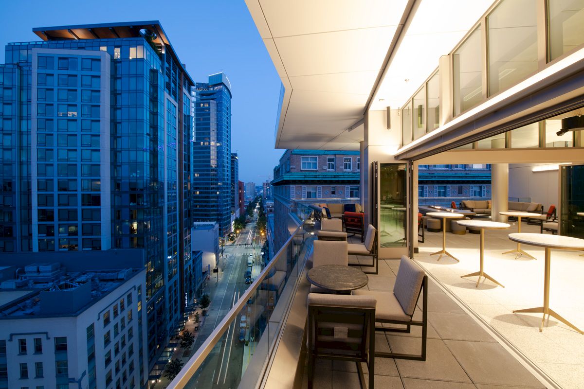 A modern rooftop balcony with seating overlooks a city street lined with tall buildings at dusk, featuring a sleek, glowing ambiance.