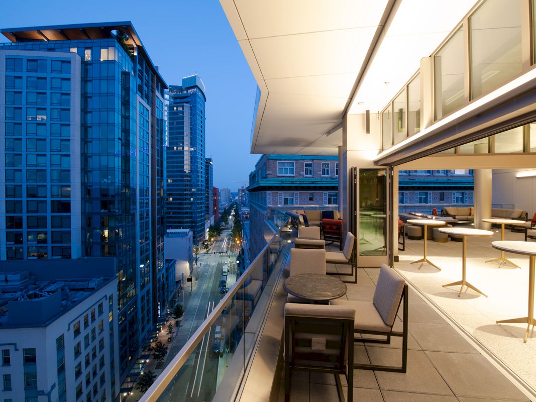 A modern rooftop balcony with seating overlooks a city street lined with tall buildings at dusk, featuring a sleek, glowing ambiance.