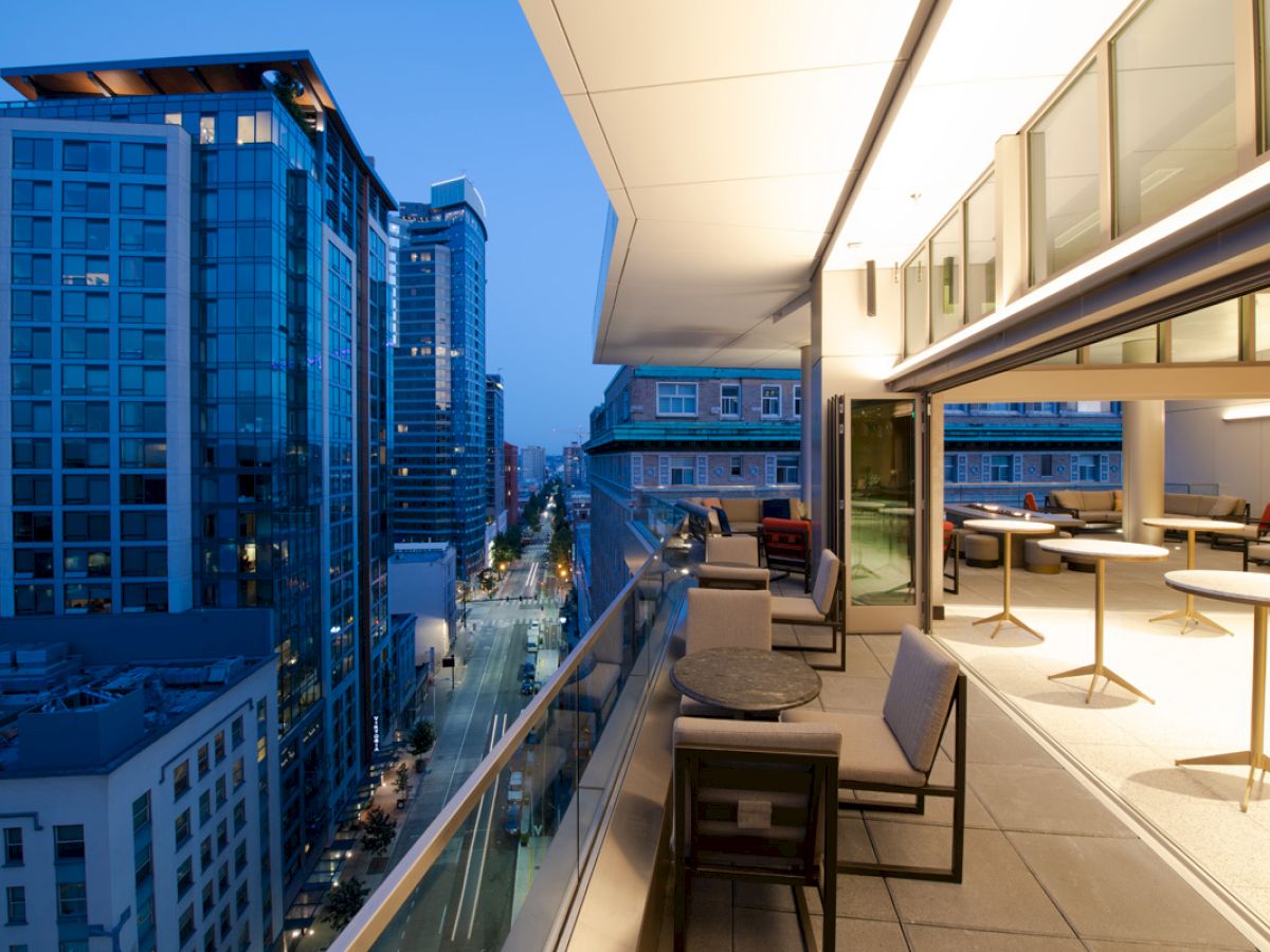 Modern rooftop terrace with tables and chairs overlooking a cityscape at dusk, surrounded by tall buildings and illuminated streets.