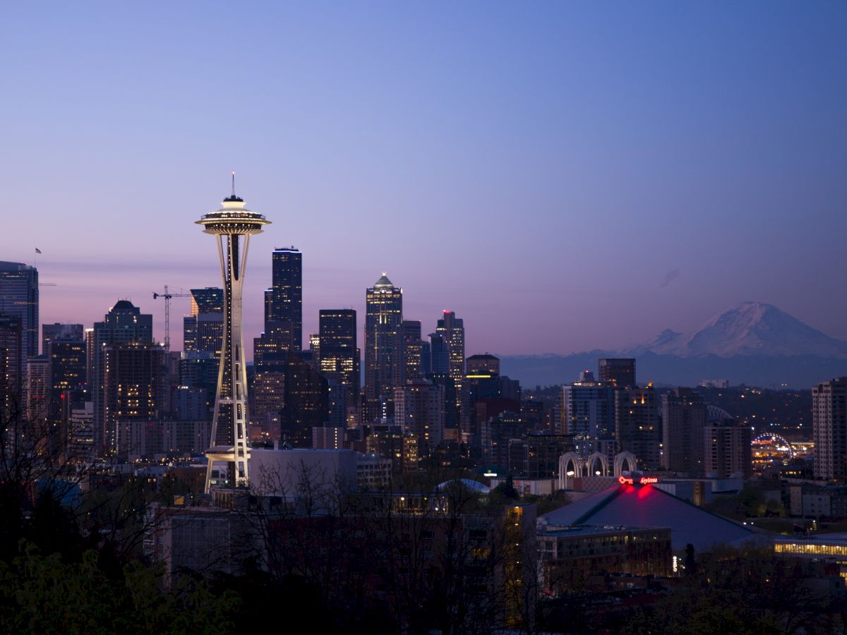 The image shows the Seattle skyline at dusk, featuring the Space Needle and Mount Rainier in the background, with city lights illuminating.