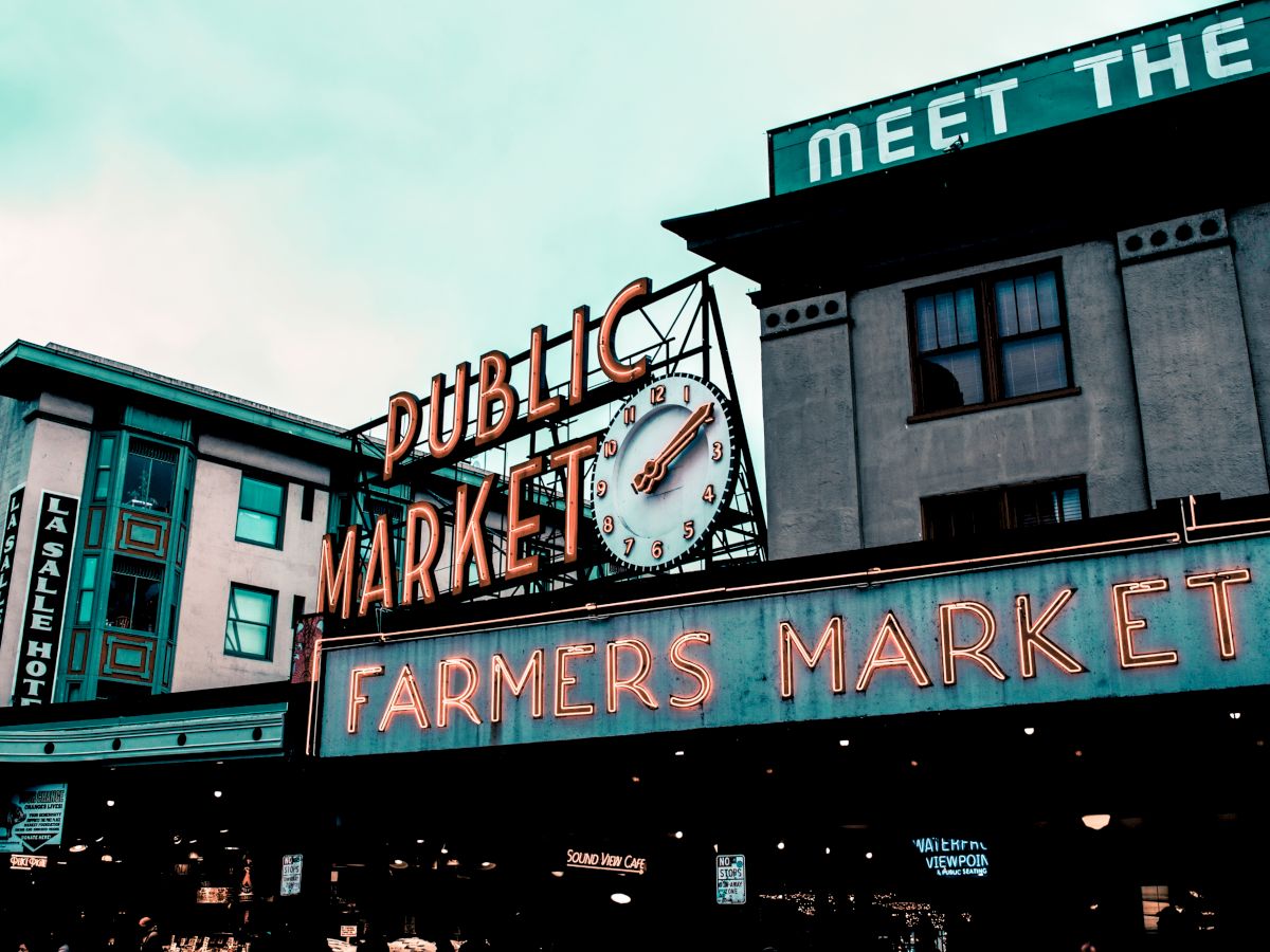 The image shows the iconic neon signs for a public market and farmers market, with a clock, against a vintage-style architectural backdrop.