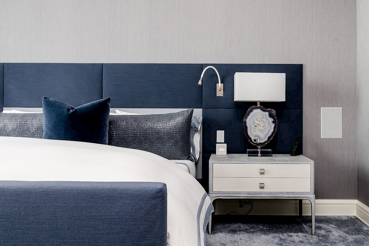 A modern bedroom with a blue and gray color scheme, featuring a bed, a side table with a lamp, and decorative pillows.