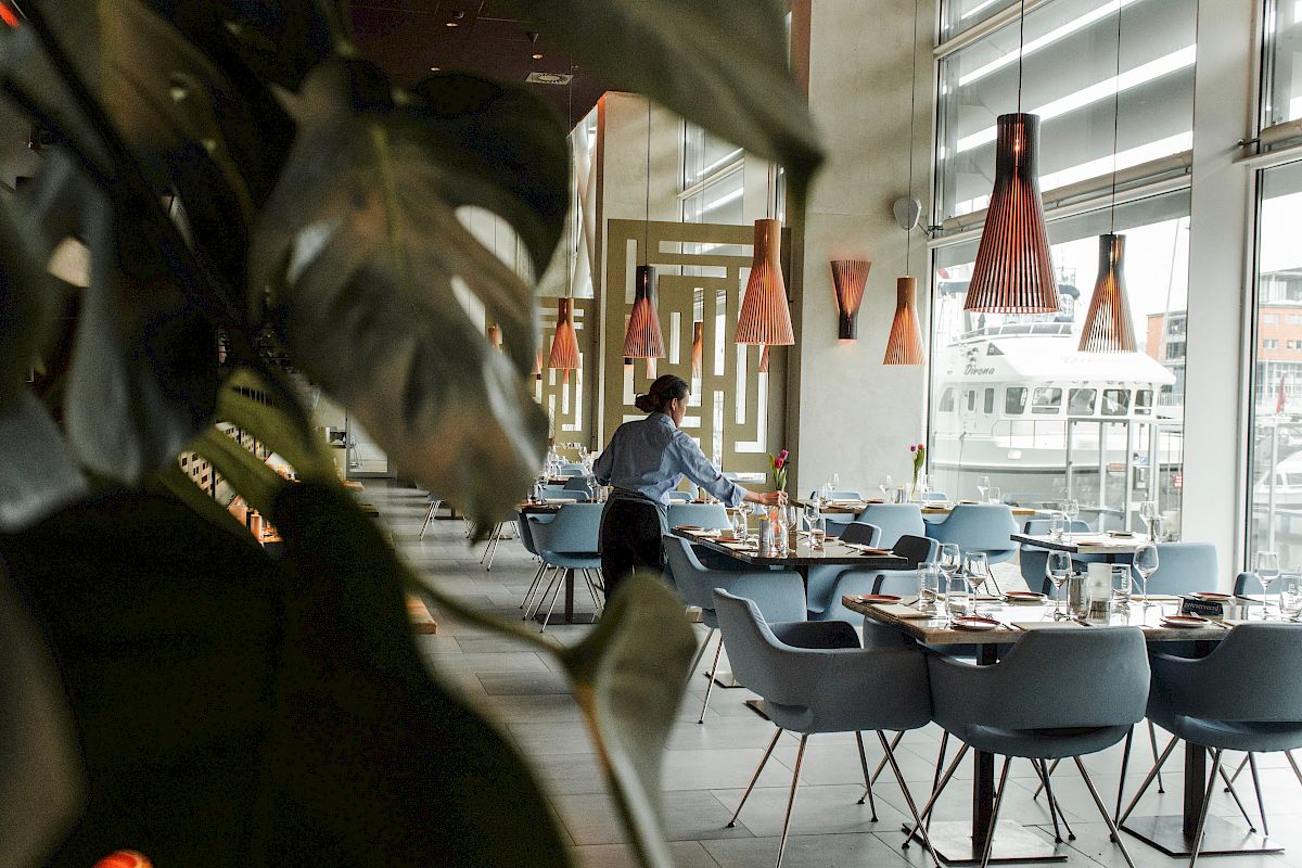 A stylish restaurant interior with a person setting tables, modern lighting, and plants in the foreground, creating a welcoming atmosphere.