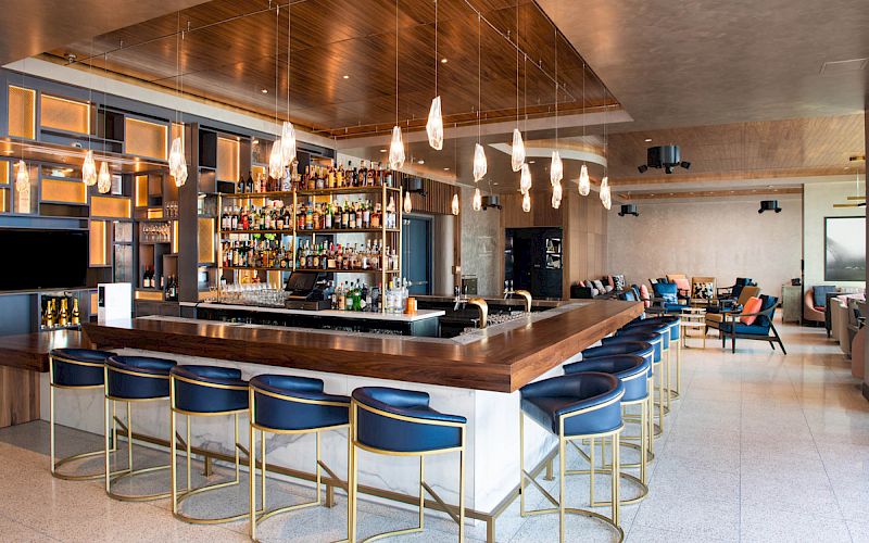 The image shows a modern bar with a wooden counter, blue chairs, hanging lights, and a well-stocked shelf. Cozy seating area in the back.