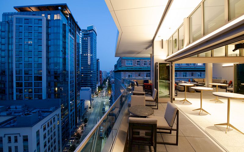 A modern cityscape at dusk with tall buildings and a stylish rooftop patio featuring seating and tables.